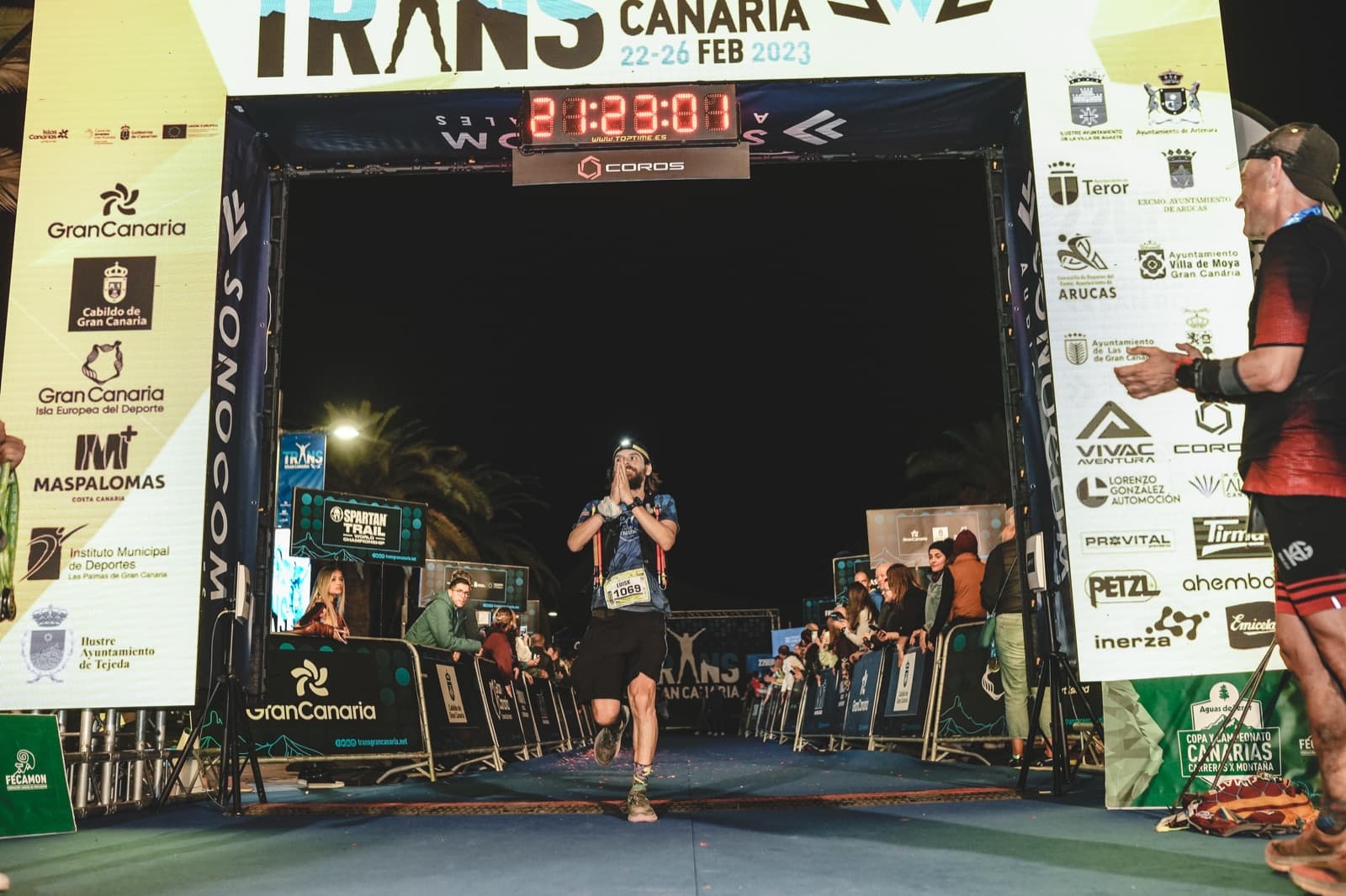 Luis crosses the finish line of Transgrancanaria 2023 at night, hands together, with a headlamp on.