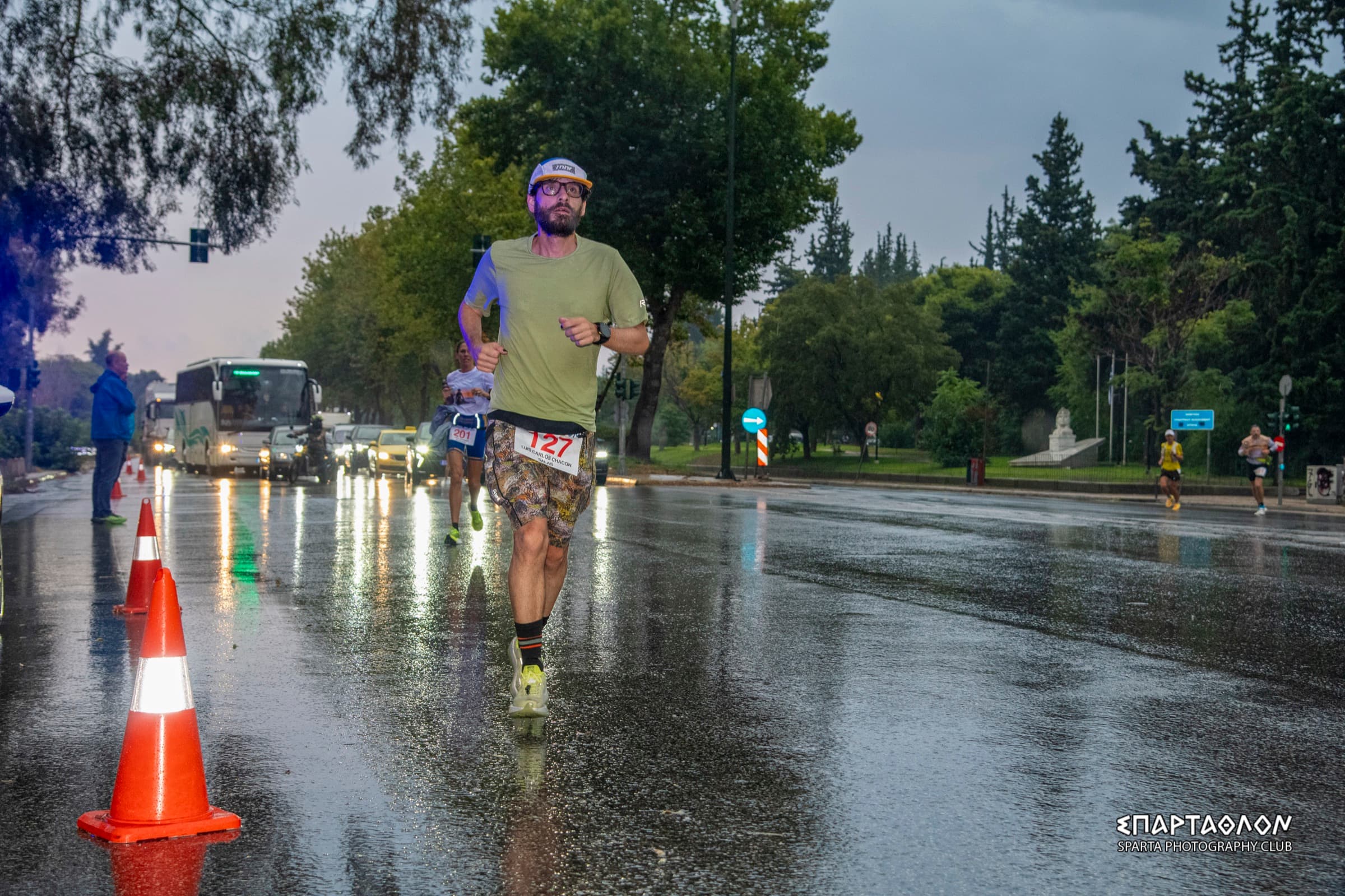 The author runs in the rain along a wet avenue in Athens, wearing bib 127, between traffic cones and other runners.