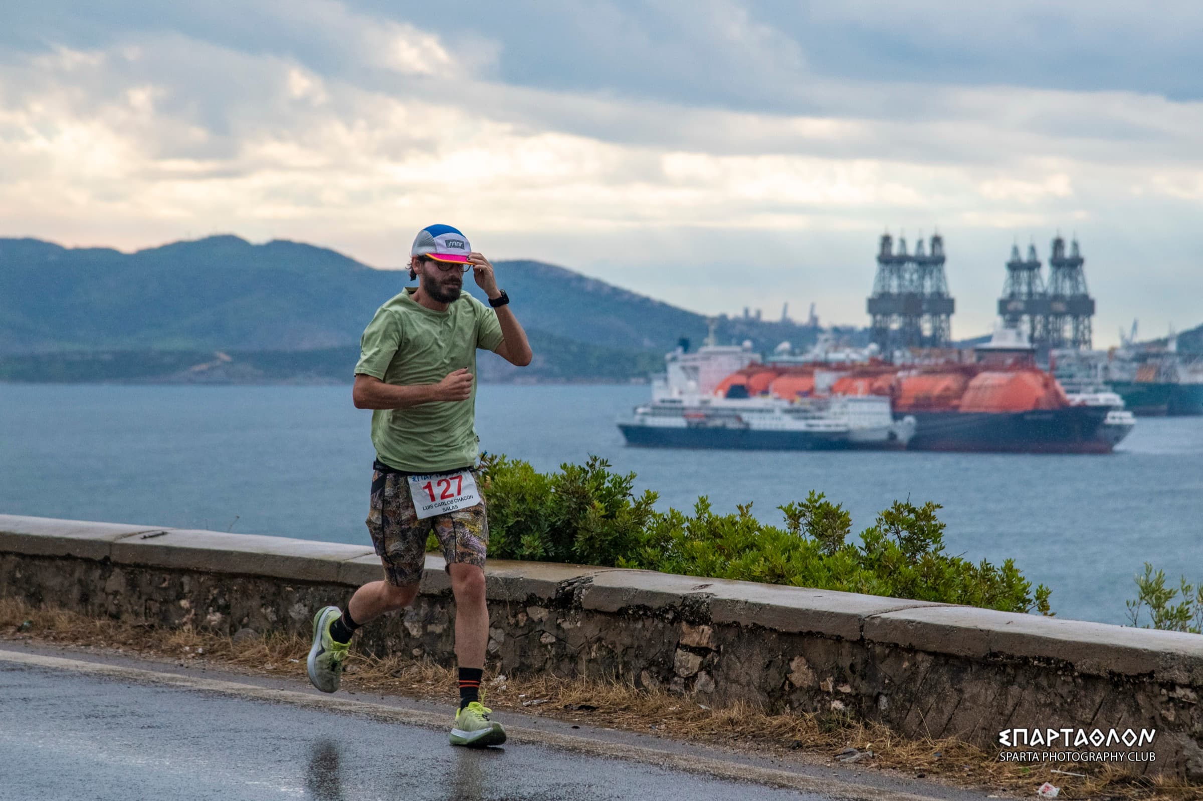 The author runs along a wet road by the sea, wearing bib 127. Behind him are large ships, shipyard cranes and mountains under a cloudy sky.