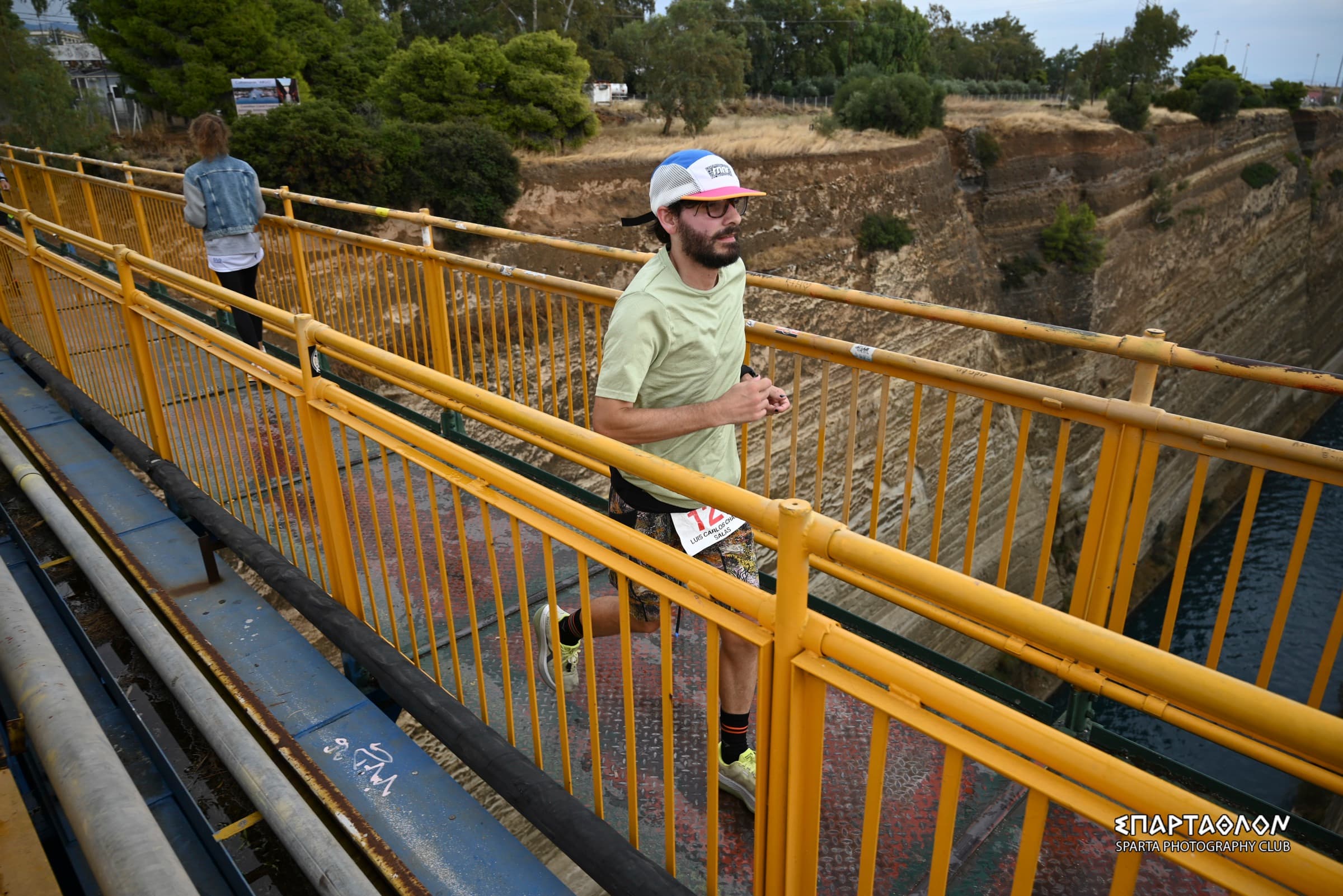 The author runs across the yellow bridge over the Corinth Canal, wearing bib 127. Below are the canal's rock walls and the water.