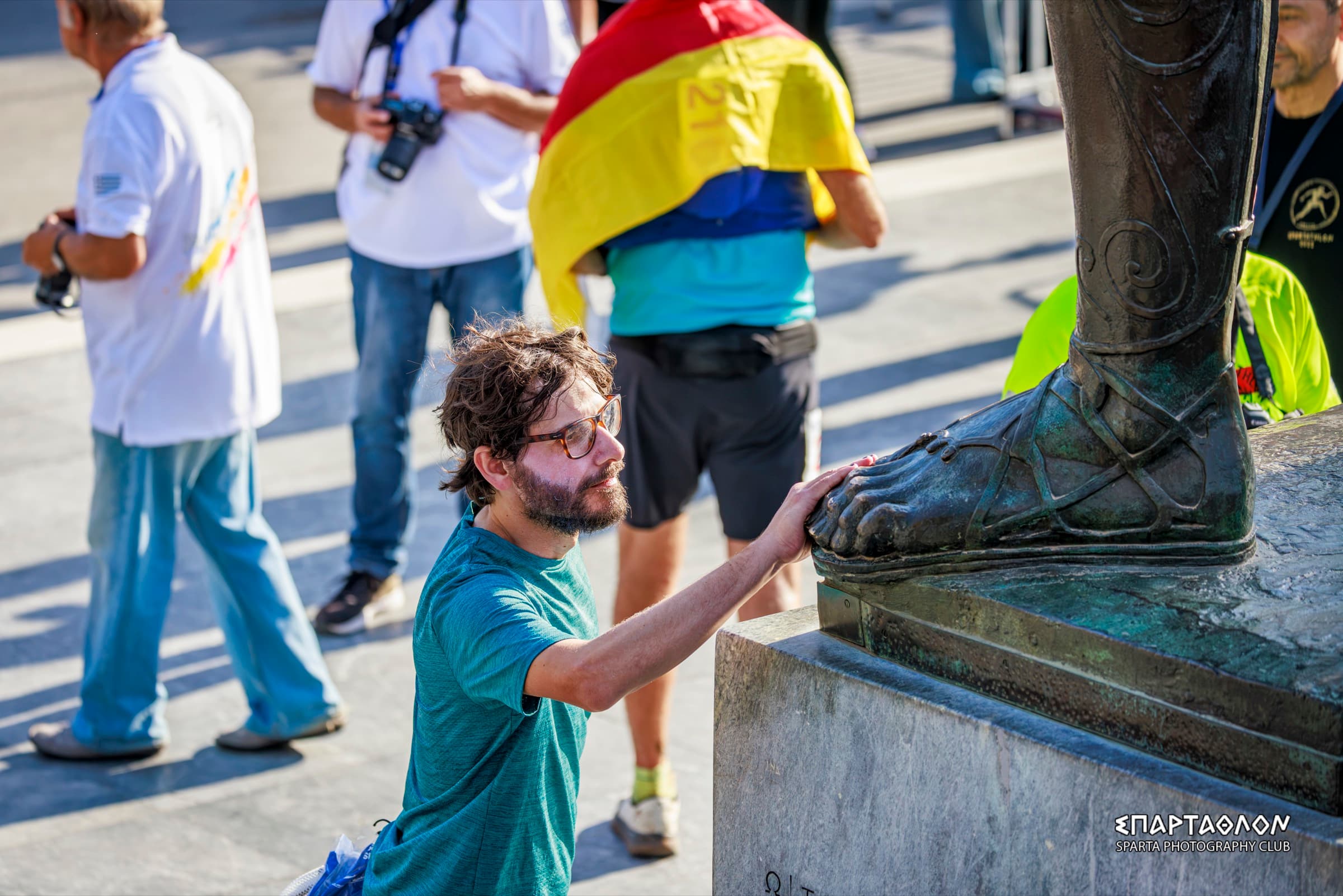 The author, kneeling by the statue of Leonidas, puts his hand on its bronze foot. Behind him are photographers and a runner wrapped in a flag.